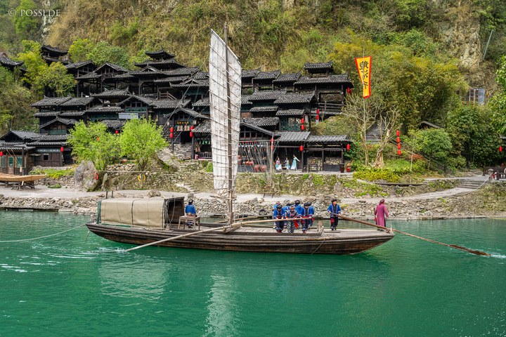 Das Bild zeigt ein traditionelles chinesisches Dorf am Ufer eines Flusses, umgeben von dichtem Grün und mit einem traditionellen Boot auf dem Wasser. (31.03.2025)
