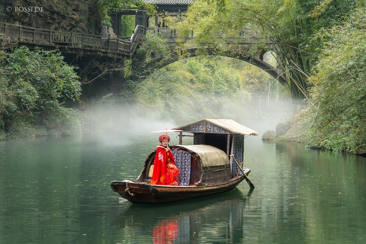 Das Bild zeigt eine idyllische Szene mit einer traditionell gekleideten Frau in einem Boot auf dem Fluss, umgeben von dichtem Grün und einer Brücke im Hintergrund. (31.03.2025)