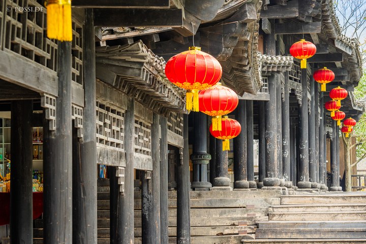 Die Fotografie zeigt eine traditionelle chinesische Architektur mit roten Laternen, die an Holzsäulen hängen und eine festliche Atmosphäre erzeugen. (31.03.2025)