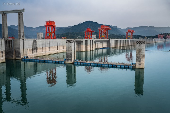 Die Aufnahme zeigt eine beeindruckende Landschaft mit einem großen Wasserreservoir, umgeben von hohen Bergen und roten Kränen. (31.03.2025)