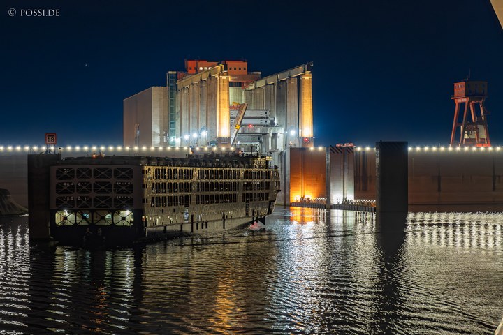 Das Bild zeigt eine Industrieanlage am Wasser, die bei Nacht beleuchtet ist und einen industriellen Charakter ausstrahlt. (31.03.2025)