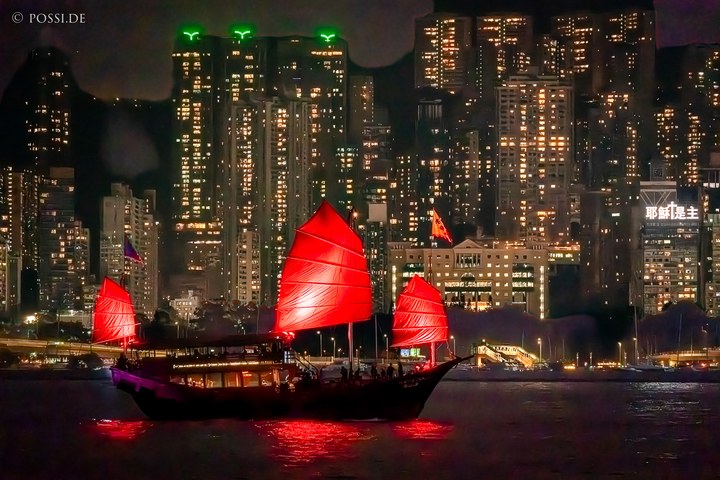 Die Szene zeigt ein beleuchtetes Boot mit roten Segeln auf einem Wasserweg vor einer beleuchteten Skyline, die wahrscheinlich Hongkong ist. (08.04.2025)